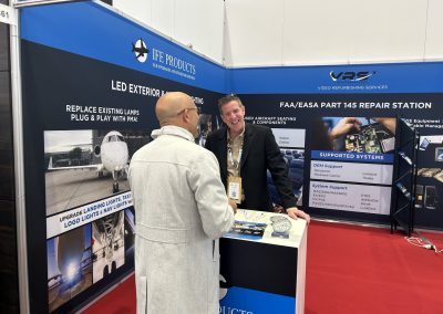 Two men talk at a trade show booth for JFE Products, featuring displays about LED exterior/interior lighting and FAA/EASA repair services. Promotional materials and aircraft components are visible on the counter.