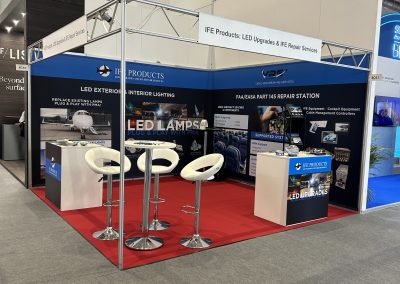 A trade show booth featuring IFE Products, with banners for LED upgrades and repair services, a counter with brochures, white stools, and a display showing aircraft lighting solutions on a red carpet.