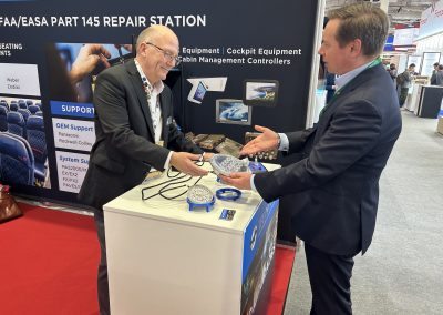 Two men in business attire stand at a trade show booth labeled FAA/EASA PART 145 REPAIR STATION, discussing aviation equipment displayed on the counter. One man gestures toward items as they converse.