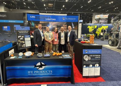 Five people stand smiling in front of an IFE Products booth at a trade show, featuring displays of LED lighting products, informational posters, bowls of oranges, and marketing materials on tables.