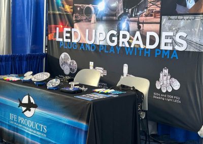 A trade show booth displays a blue IFE Products tablecloth, LED lights on the table, colorful cards, and a large backdrop reading LED UPGRADES PLUG AND PLAY WITH PMA beside aircraft and LED product images.