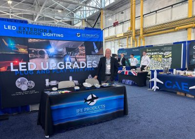 A man stands at an exhibition booth for IFE Products, displaying LED lighting upgrades. Other booths and people are visible in the background, with banners, displays, and aviation-themed items present.